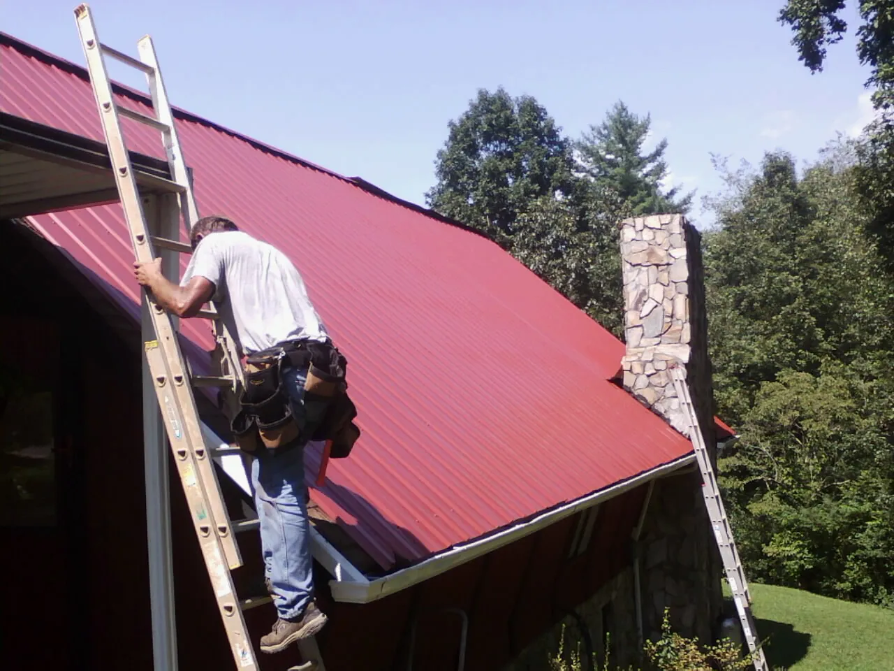 Roofer on a ladder beside a red metal roof and stone chimney — Soffit & Fascia Repair by Roofing Brothers in Lebec